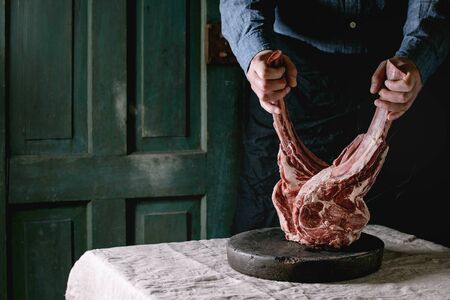 Man's hands holding raw uncooked black angus beef tomahawk steaks on bones on linen table cloth....