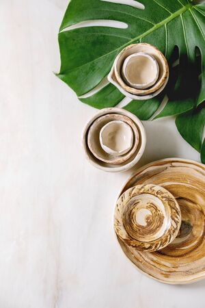 Variety of empty different ceramic dishes, bowls, jugs and plates on exotic monstera leaves over white marble background. Flat lay, spaceの写真素材