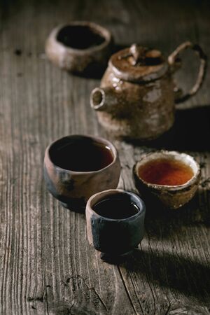 Hot black tea in different traditional wabi sabi style fireclay ceramic craft cups served with plate, teapot and bowl of dry tea leaves on old wooden table. Dark rustic atmosphere.の写真素材