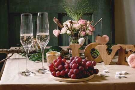 st. Valentines day or wedding romantic table setting with wooden letters love, needle felted hearts, bouquet of pink roses, couple glasses of champagne and red grapes on natural linen tablecloth.の写真素材