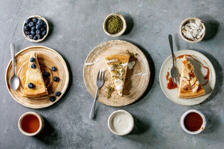 Pieces of cheesecake with different toppings on ceramic plates and variety of sauces in ceramic bowls in row over grey texture background. Flat lay, spaceの写真素材