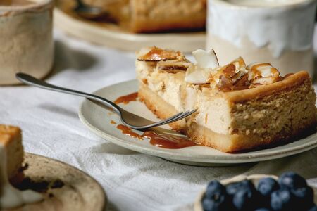 Piece of cheesecake with caramelized coconut chips and caramel topping on ceramic plates on white...
