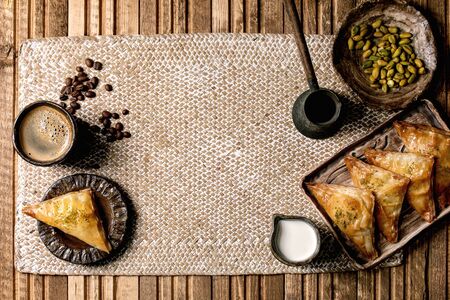 Homemade Turkish traditional dessert baklava with pistachio served on ceramic plate with bowl of nuts, cup of coffee, cream, cezva on straw napkin over wooden background. Flat lay, spaceの写真素材