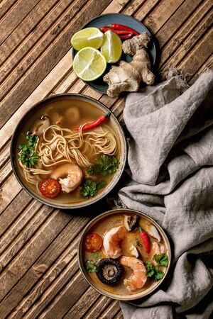 Variety of asian soups. Traditional spicy Thai tom yum kung and noodles soup with shiitake mushrooms, prawns, ingredients above on linen cloth over wooden plank background. Flat lay, space.の写真素材