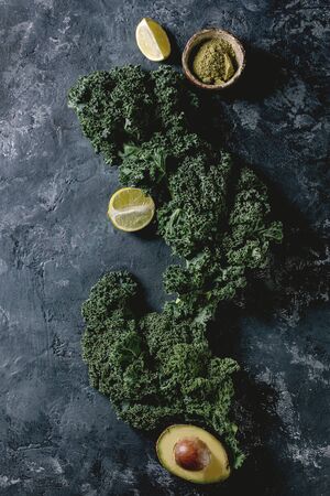 Ingredients for smoothie. Heap of fresh organic curly kale leaves, half of avocado, limes, matcha powder over black concrete background. Healthy eating ingredients. Top view, copy spaceの写真素材