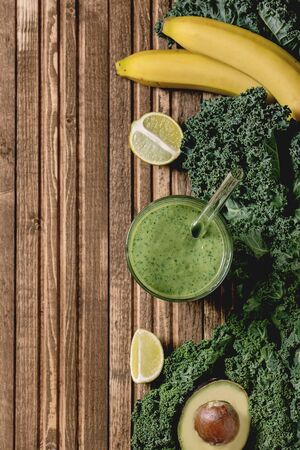 Glass of green healthy vegan smoothie, glass straw. Ingredients above. Kale, bananas, avocado, lime, over wooden plank background. Flat lay, spaceの写真素材