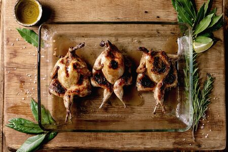 Roasted grilled butterfly quails in glass baking tray with greens, salt and olive oil over wooden background. Flat lay, spaceの写真素材