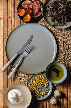Ingredients for cooking salad. Italian burrata cheese, black arugula, grilled apricot, prosciutto, pistachio nuts and olive oil in bowls on round knitted napkin over wooden background. Flat lay, spaceの写真素材