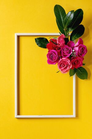 Creative layout with beautiful pink roses flowers and green leaves in wooden frame on bright yellow background. Flat lay. Greeting card conceptの写真素材