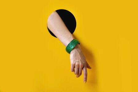 Female hand with yellow manicure and plastic green bracelet points or touch to something from black round hole on yellow background. Trendの写真素材