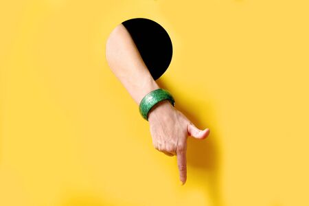 Female hand with yellow manicure and plastic green bracelet points to something from black round hole on yellow background. Trendの写真素材