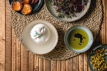 Ingredients for cooking salad. Italian burrata cheese, black arugula, grilled apricot, prosciutto, pistachio nuts and olive oil in bowls on round knitted napkin over wooden background. Flat lay, spaceの写真素材