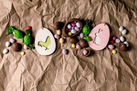 Easter greeting card with wooden rabbit and chicken decorations, chocolate sweets and eggs on crumpled craft paper background. Flat lay, copy space.の写真素材