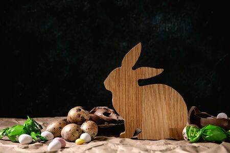 Easter mood still life with wooden rabbit decoration, golden quail eggs, traditional chocolate eggs and sweets on table with crumpled craft paper. Dark background.の写真素材