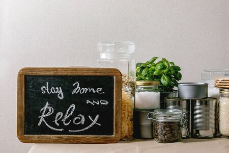 Food supplies crisis food stock for quarantine isolation period. Different glass jars with grains, pasta, cans of canned food, toilet paper, chalkboard handwritten chalk lettering Stay home and relax.の写真素材