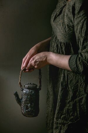 Woman in dark dress hold in hands craft handmade ceramic teapot with twig wooden handle for serving tea drinking.の写真素材