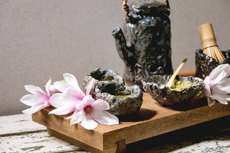 Japanese matcha green tea in craft ceramic cups with matcha powder, bamboo whisk, ceramic teapot and pink magnolia flowers on traditional wooden japanese table.の写真素材