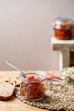 Sun-dried cherry tomatoes olive oil in glass jar with fork, standing on wooden kitchen table with sliced rye bread and green sprouts salad.の写真素材