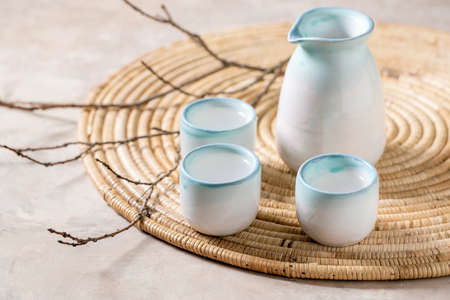 Sake ceramic set for traditional japanese alcohol drink rice wine sake, pitcher and three cups, standing on straw napkin with dry branches over beige texture background.の写真素材