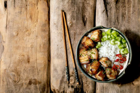 Grilled deep fried pork belly in bowl with rice, celery, chili pepper and spring onion with chopsticks over old wooden background. Flat lay, copy spaceの写真素材