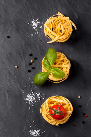 Top view on dry pasta, fresh tomatoes, yellow chili pepper and basil served with sea salt on dark gray backgroundの写真素材