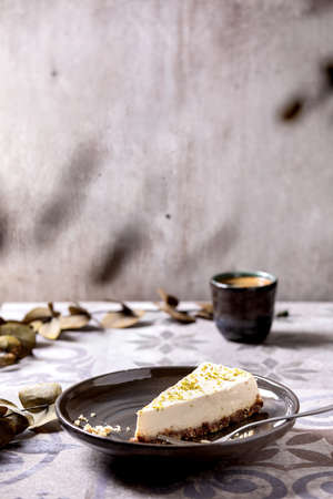 Piece of raw vegan cheesecake, no bake gluten free, decorated by lime zest and cashew nuts on plate. Cup of black coffee and Eucalyptus branches on ornate ceramic table. Sunlightの写真素材