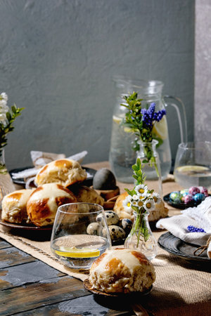 Easter table setting with colored and chocolate eggs, hot cross buns, bouquet flowers, empty ceramic plate with napkin, glass of lemonade drink on wooden table with textile tablecloth.の写真素材