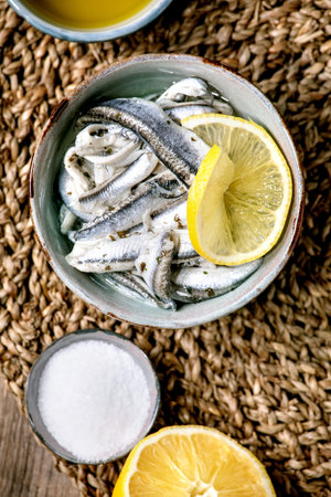 Bowl of pickled anchovies or sardines fillet in oil, served with lemon, olive oil, salt for tapas or antipasti over old wooden background. Flat lay, copy spaceの写真素材