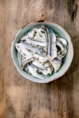 Bowl of pickled anchovies or sardines fillet in oil with herbs for tapas or antipasti over old wooden background. Flat lay, copy spaceの写真素材