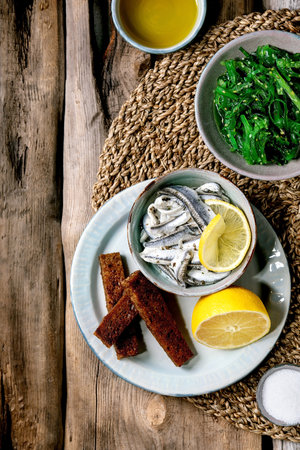Bowl of pickled anchovies or sardines fillet in oil, served with lemon, olive oil, salt and toasted rye bread for tapas or antipasti, Wakame seaweed salad on old wooden background. Flat lay.の写真素材
