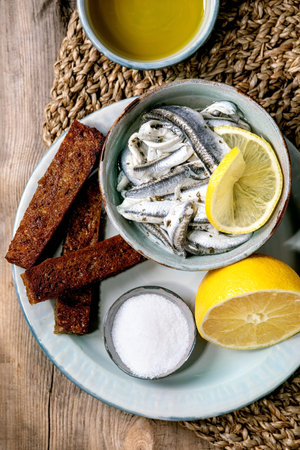 Bowl of pickled anchovies or sardines fillet in oil, served with lemon, olive oil, salt and toasted rye bread for tapas or antipasti over old wooden background. Flat lay, copy spaceの写真素材