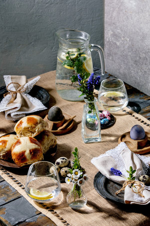 Easter table setting with colored and chocolate eggs, hot cross buns, bouquet flowers, empty ceramic...
