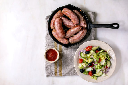 Grilled italian sausages salsiccia in cast-iron pan served with tomato sauce and plate of fresh vegetable salad on white marble background. Balanced dinner. Flat lay, copy spaceの写真素材