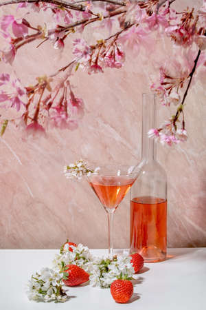 Concept of strawberry alcoholic or non-alcohol cocktail in martini glass, and glass bottle decorated with blooming branches of cherry tree over white and pink marble backgroundの写真素材