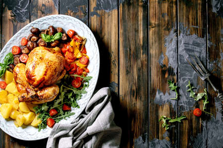 Grilled cooked whole chicken with vegetable garnish grilled bell pepper, onion, baked potatoes, cherry tomatoes, mushrooms, herbs in ceramic plate over dark wooden background. Flat lay. Copy spaceの写真素材