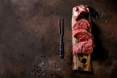 Fresh sliced raw beef tenderloin meat for steaks on wooden board with metal meat fork, salt, pepper...