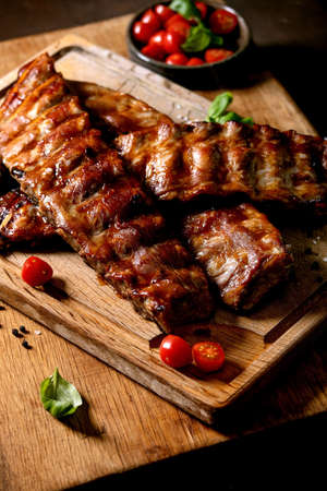 Grilled pork bbq ribs served with cherry tomatoes, basil and barbeque sauce on wooden cutting board over dark background. Close upの写真素材