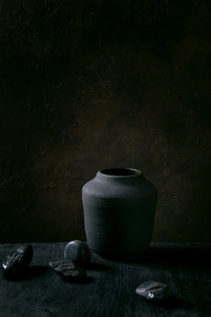 Empty black ceramic vase on black wooden table with decorative stones. Dark still life. Copy space.の写真素材