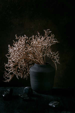 Dry white flowers branch in black ceramic vase on black wooden table with decorative stones. Dark...