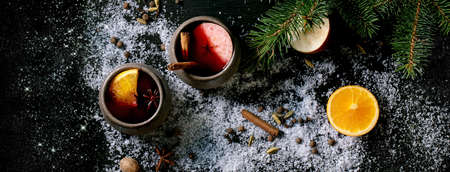 Hot mulled wine in two ceramic mugs with spices, orange, apple slice and Christmas entourage over black wooden background with fir tree and snow. Flat lay, banner sizeの写真素材