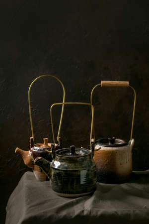 Variety of three craft handmade ceramic teapots kettles for tea ceremony standing on linen tablecloth in dark room.の写真素材