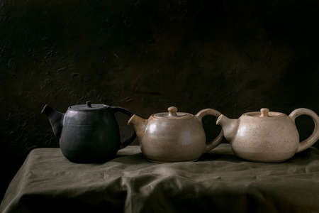 Variety of craft handmade ceramic teapots kettles in row for tea ceremony standing on linen tablecloth in dark room.の写真素材