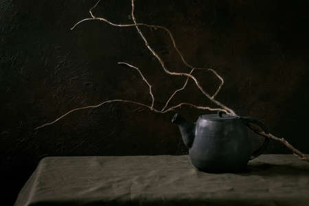 Caft handmade black ceramic teapot kettle for tea ceremony standing on linen tablecloth with dry branch in dark room.の写真素材