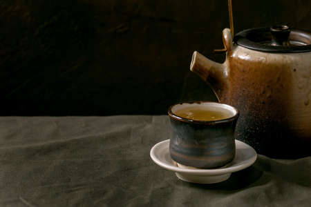 Craft handmade ceramic teapot kettle with cup of hot green tea with saucer, standing on linen tablecloth in dark room.の写真素材