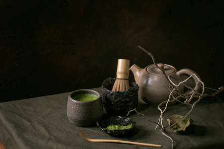 Traditional japanese hot green frothy tea matcha in ceramic cup, powdered matcha, teapot and bamboo whisk standing on dark linen tablecloth. Healthy drink. Dry branch and leaf as decor.の写真素材