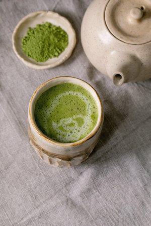 Traditional japanese hot green frothy tea matcha latte in handmade ceramic cup, powdered matcha, teapot and bamboo whisk standing on grey linen tablecloth. Healthy drinkの写真素材