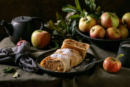 Homemade sliced traditional austrian apple strudel pie in black ceramic plate served with ripe fresh apples, branches, fork and icing sugar on dark linen tablecloth. Dark rustic Autumn mood.の写真素材