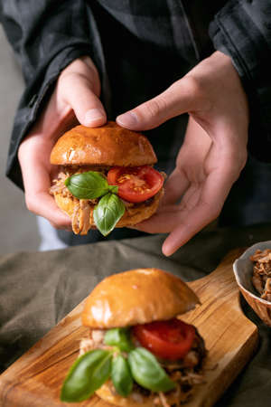 Young man chef hold in hand mini burger with stew beef, tomatoes and basil over kitchen table with other hamburgers. Modern delicious fast foodの写真素材