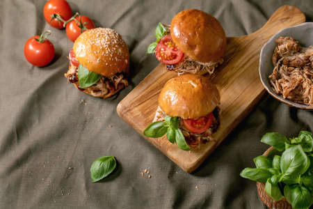 Set of homemade mini burgers with stew beef, tomatoes and basil on wooden chopping board over linen tablecloth as background. Crumbs and ingredients around. Modern delicious fast foodの写真素材