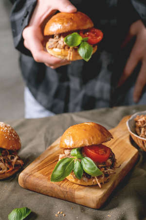 Young man chef hold in hand mini hamburger with stew beef, tomatoes and basil over kitchen table with other burgers. Modern delicious fast foodの写真素材
